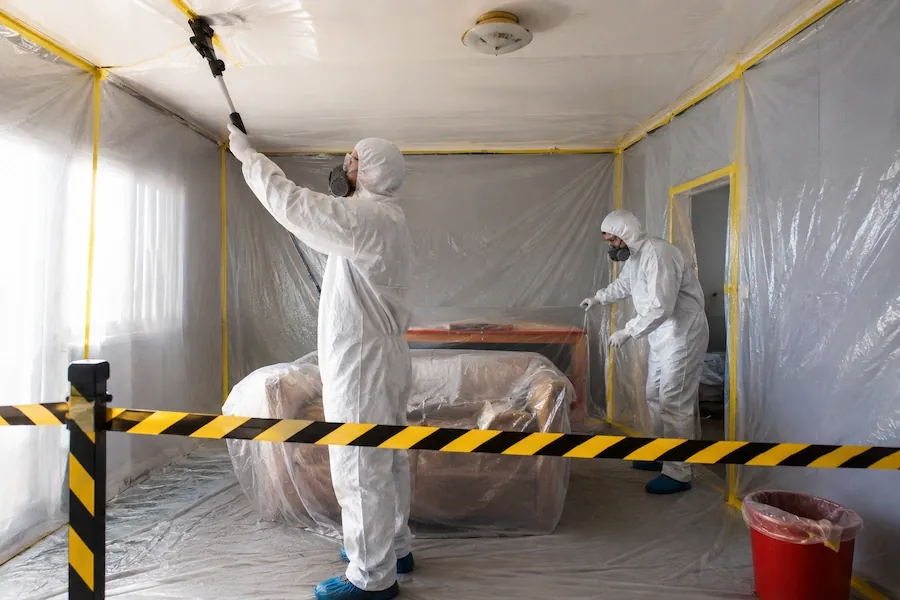 Two people in white protective suits and masks removing a ceiling inside a room sealed with plastic sheeting