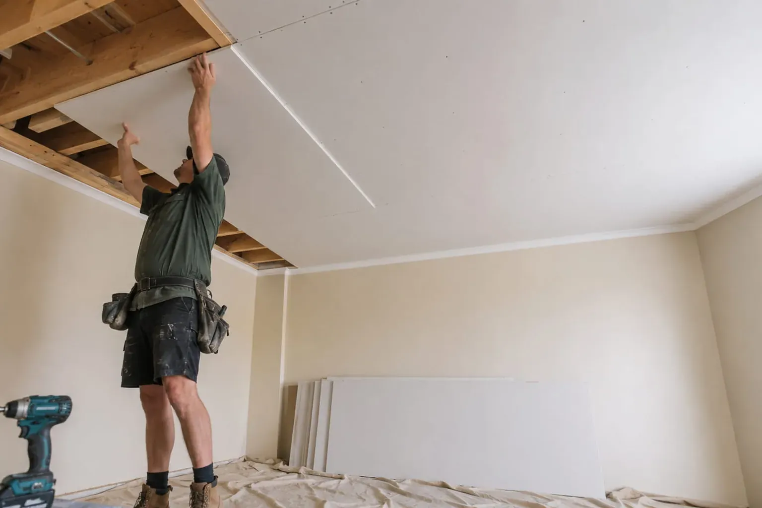 Ceiling fixer lifting a plasterboard sheet up to the timber joists, drill on the floor beside him
