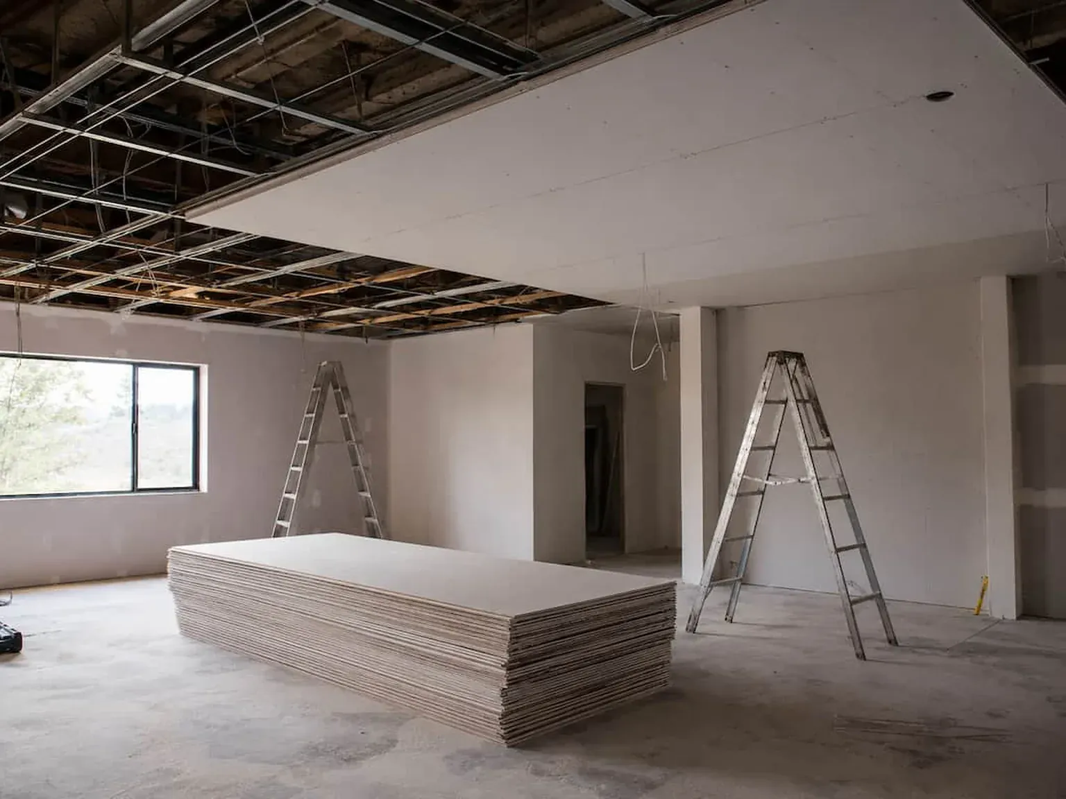 Stack of plasterboard sheets on the floor of an empty room with two ladders and a furring-channel ceiling half sheeted