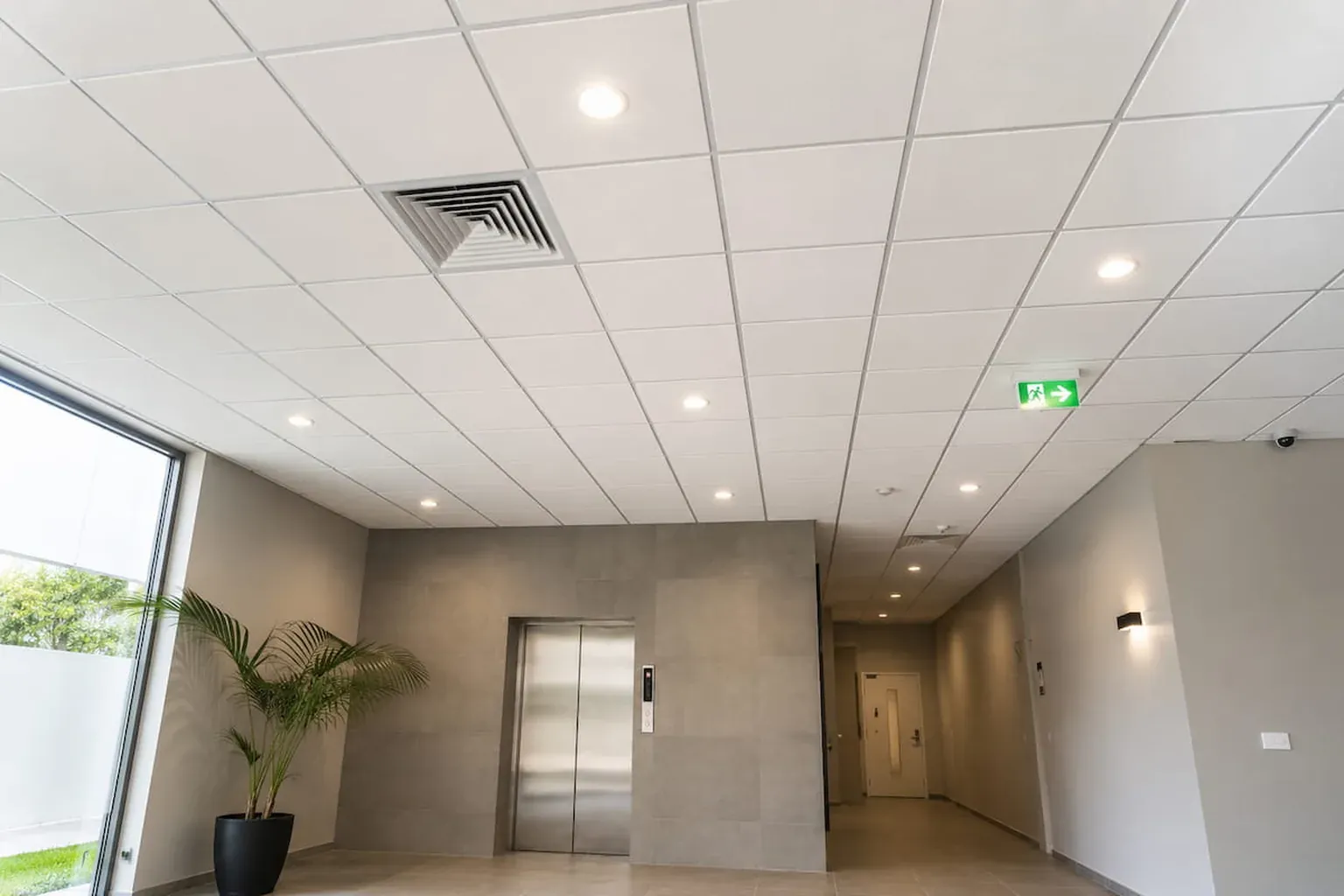 Suspended grid ceiling with downlights and an air vent above an office lobby and lift